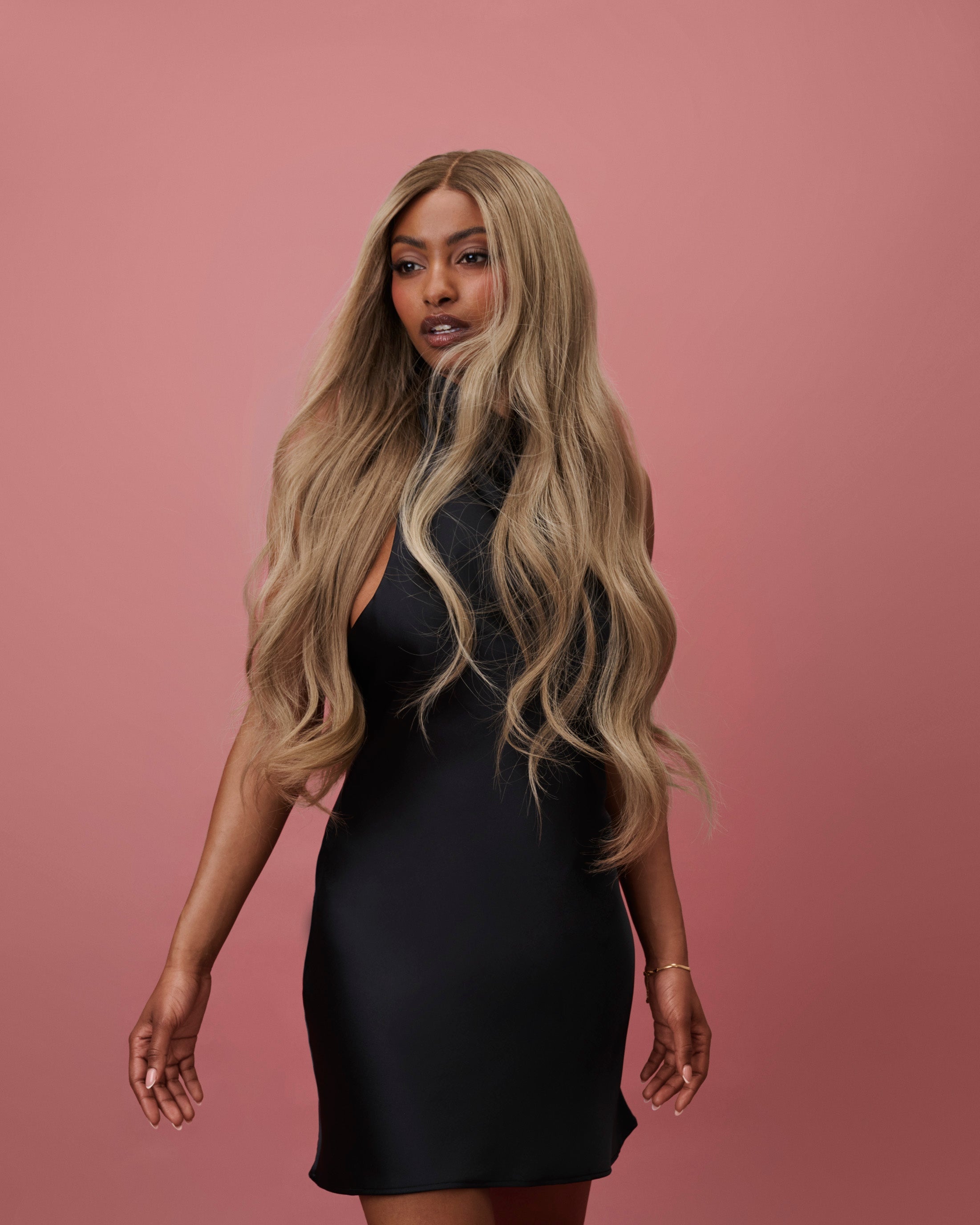 BHBD Lana Wig 65 cm – cool caramel blonde human hair wig with darker roots and softly layered 65 cm length, pre-plucked hairline and bleached knots for a natural scalp look, shown on a model wearing a black dress against a pink background