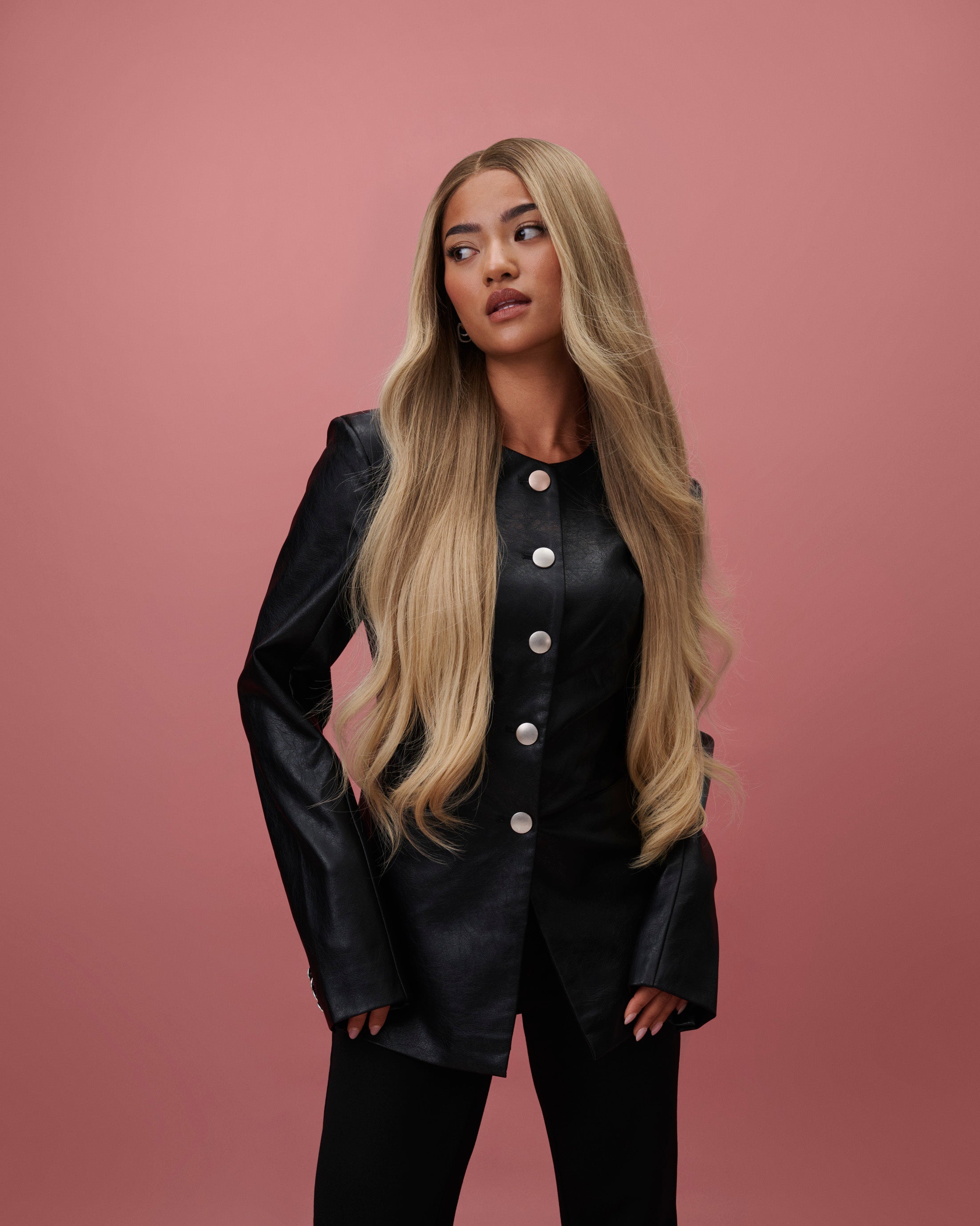BHBD Mila Wig 65 cm – cool medium blonde human hair wig that transitions into a light warm blonde length with softly layered 65 cm length, pre‑plucked hairline and bleached knots, shown on a model wearing a black outfit against a pink background