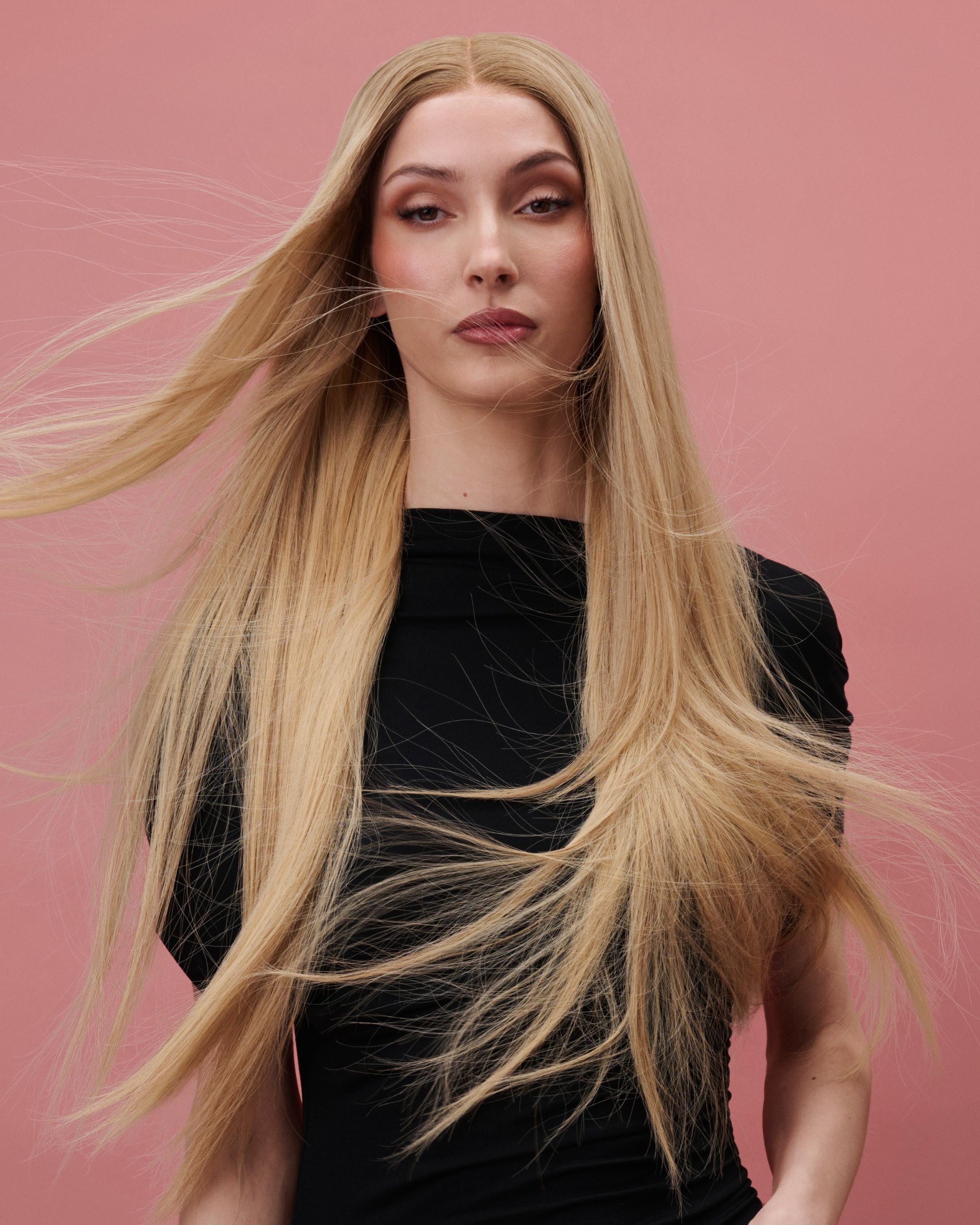 Vera Wig / 65cm – 65 cm light blonde Remy human hair wig with golden undertones and darker roots, displayed on a model facing forward against a pink background.