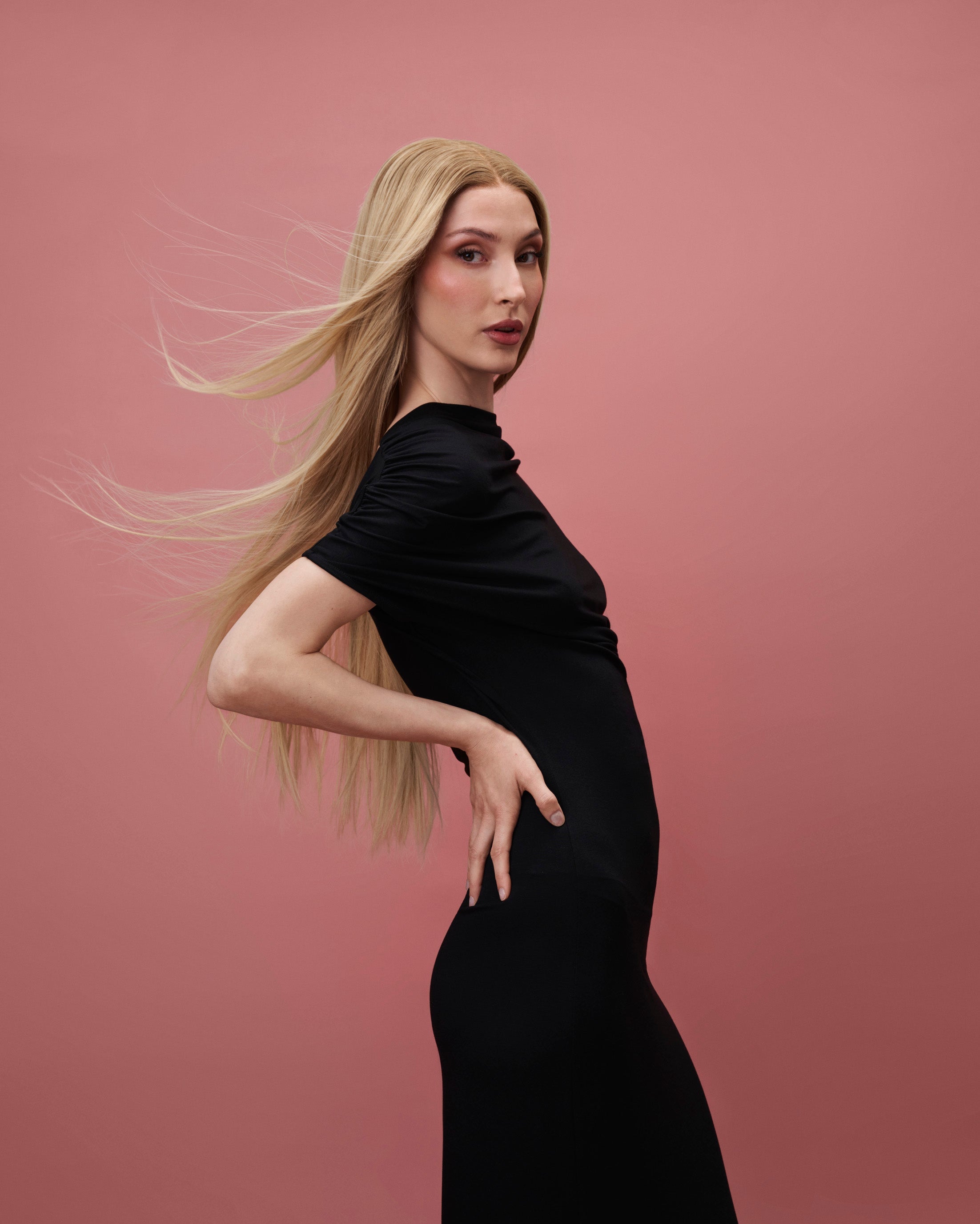 Vera Wig / 65cm – 65 cm light blonde Remy human hair wig with subtle golden tones and darker roots, softly layered, shown on a model in a black dress against a pink background.