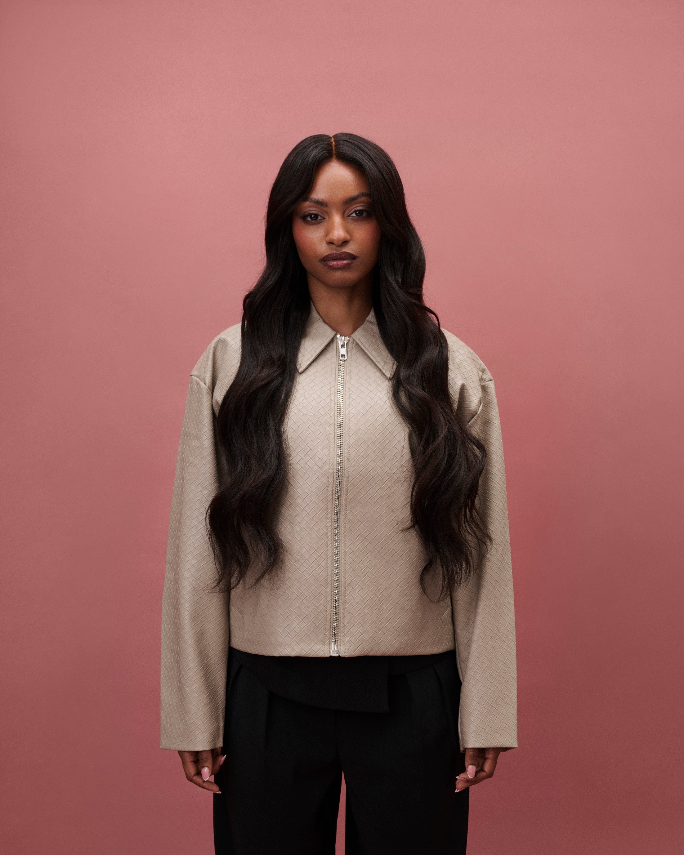 Aurora Wavy Wig 65 cm – deep natural brown human hair wig with subtle warmth and softly permed beach waves on HD lace featuring a pre‑plucked hairline and bleached knots, worn by a model in a beige top and black pants against a pink background