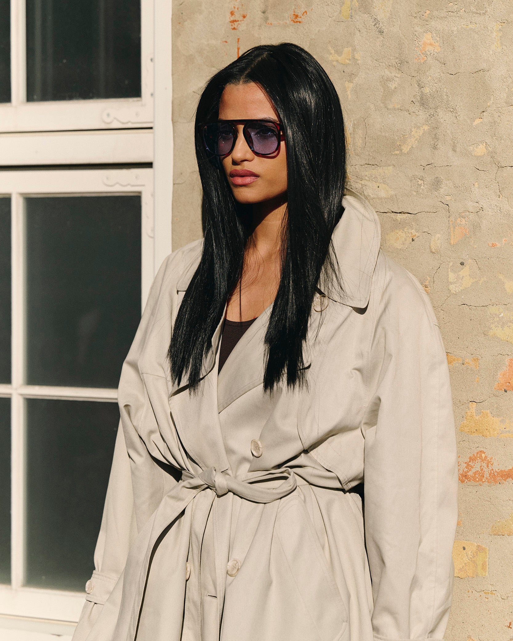 Woman in a black wig wearing a beige trench coat and sunglasses against a stone wall.