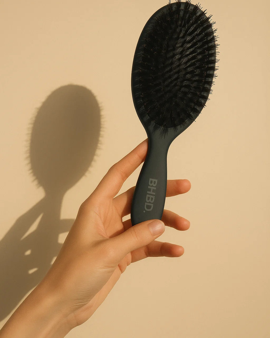BHBD Paddle Brush – hand holding a black paddle hairbrush with natural boar and nylon bristles for smoothing and detangling hair against a beige background