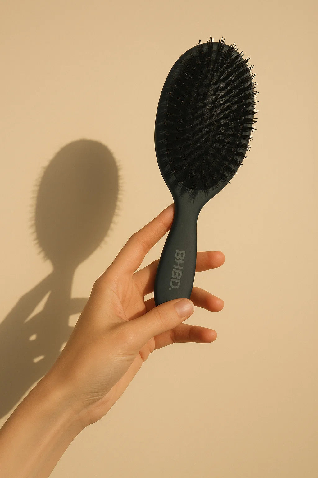 BHBD Paddle Brush – hand holding a black paddle hairbrush with natural boar and nylon bristles for smoothing and detangling hair against a beige background