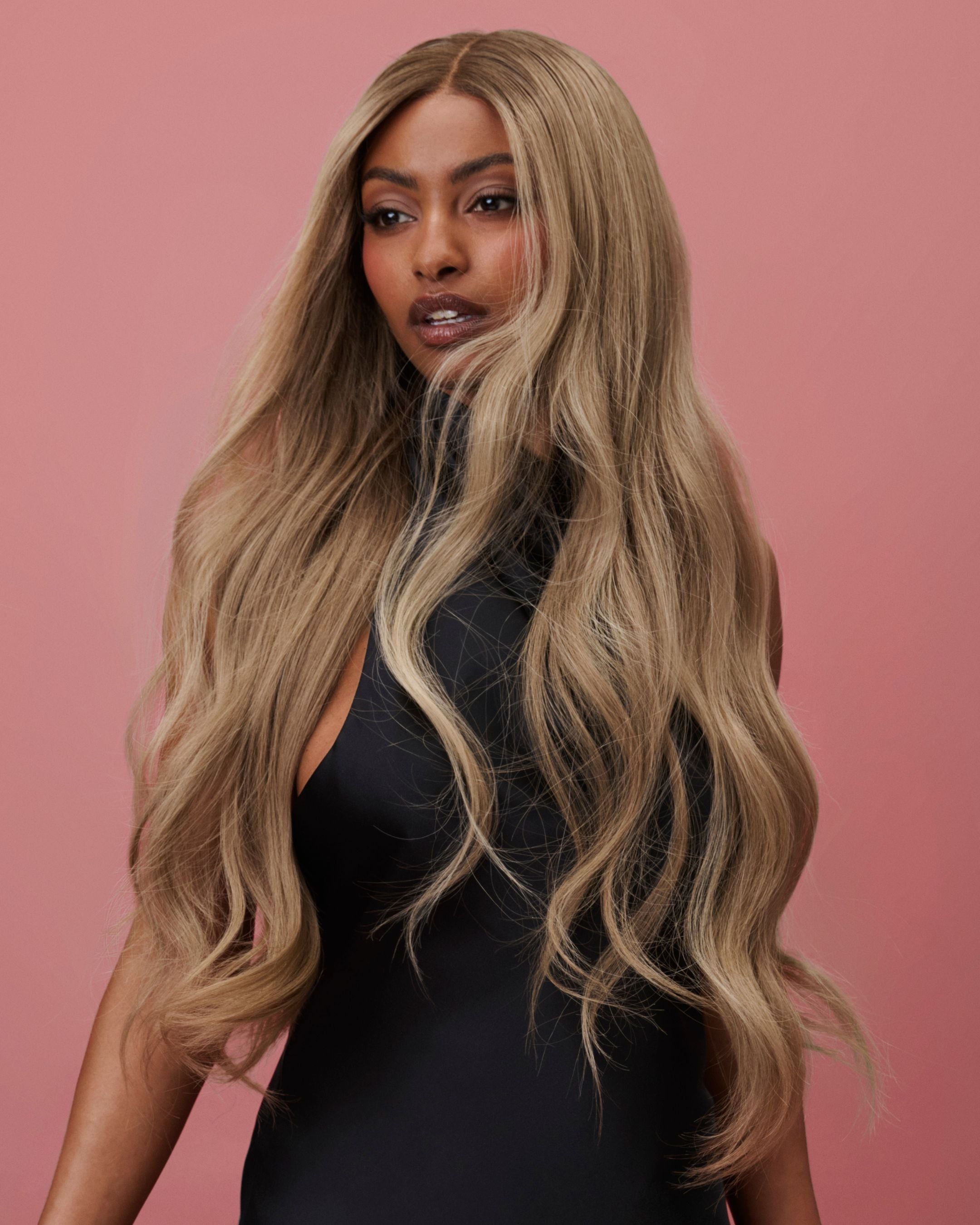 BHBD Lana Wig 65 cm – cool caramel blonde human hair wig with darker roots and softly layered 65 cm length, pre-plucked hairline and bleached knots for a natural scalp look, shown on a model wearing a black dress against a pink background