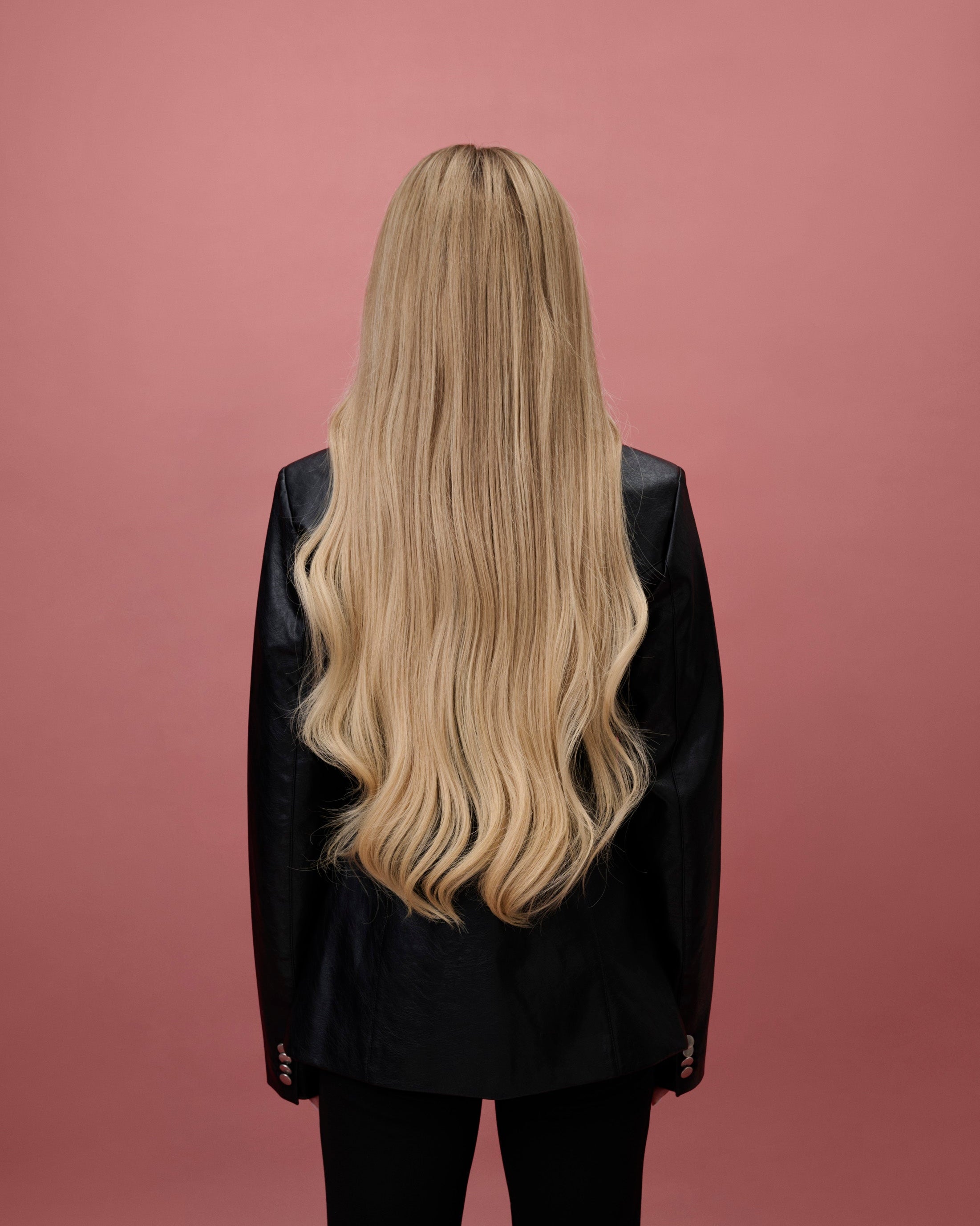 BHBD Mila Wig 65 cm – back view of cool medium blonde human hair wig transitioning to a light warm blonde length with softly layered 65 cm length and pre‑plucked hairline, shown on a model wearing a black jacket against a pink background