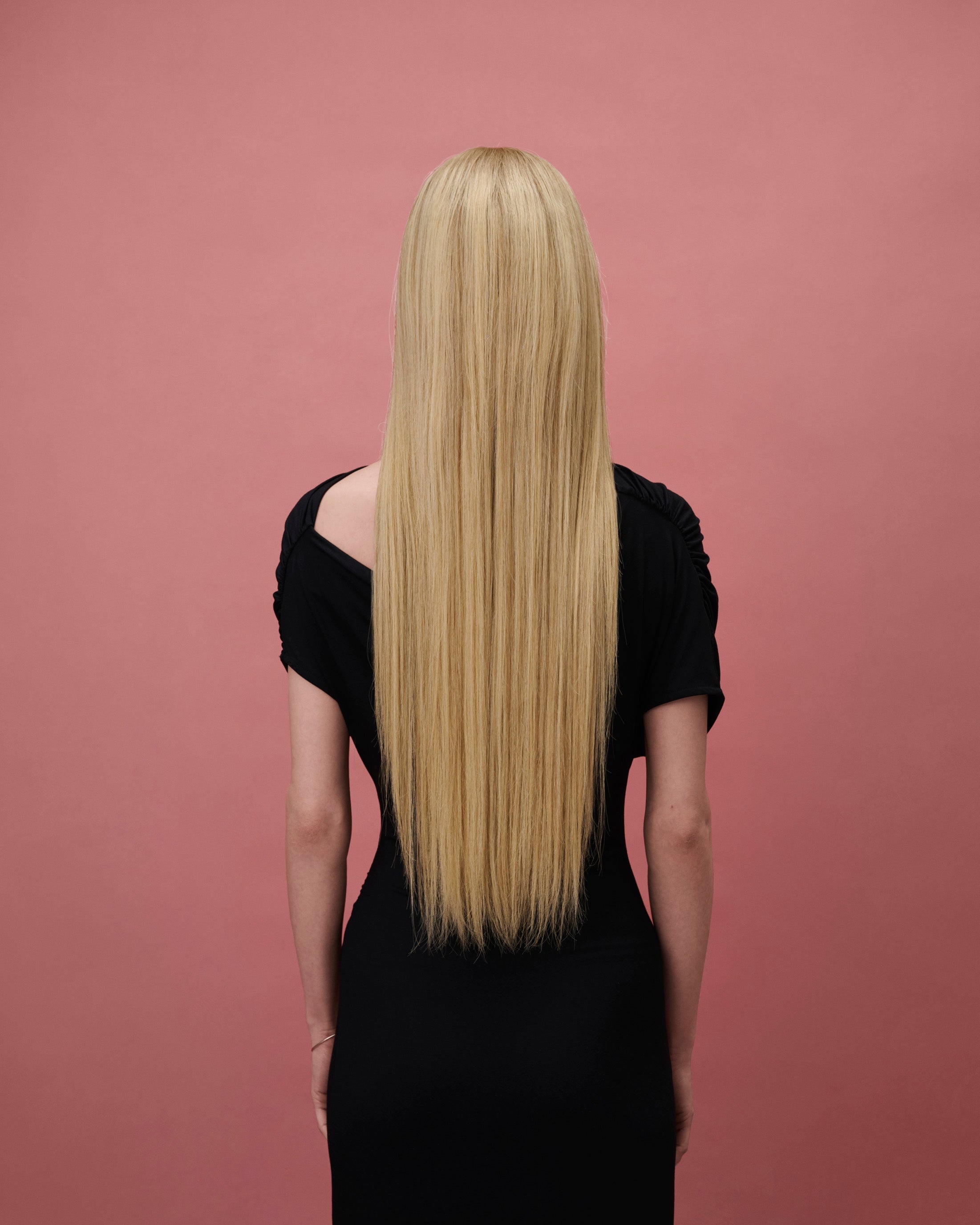 Vera Wig / 65cm – 65 cm light blonde Remy human hair wig with darker roots, straight and sleek, shown from the back on a model against a pink background.