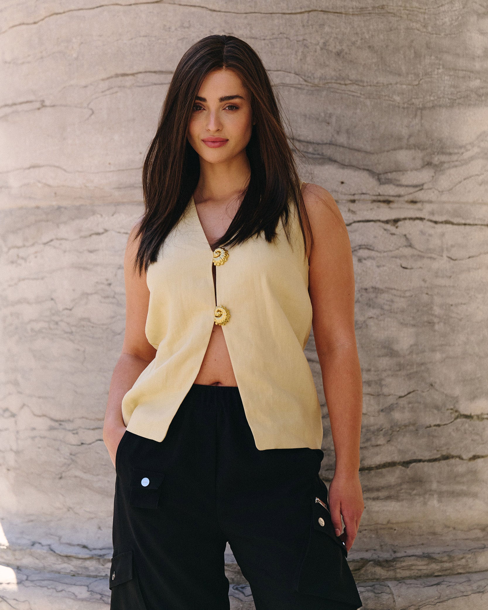 Woman wearing dark brown wig and a yellow sleeveless top with buttons against a stone wall.
