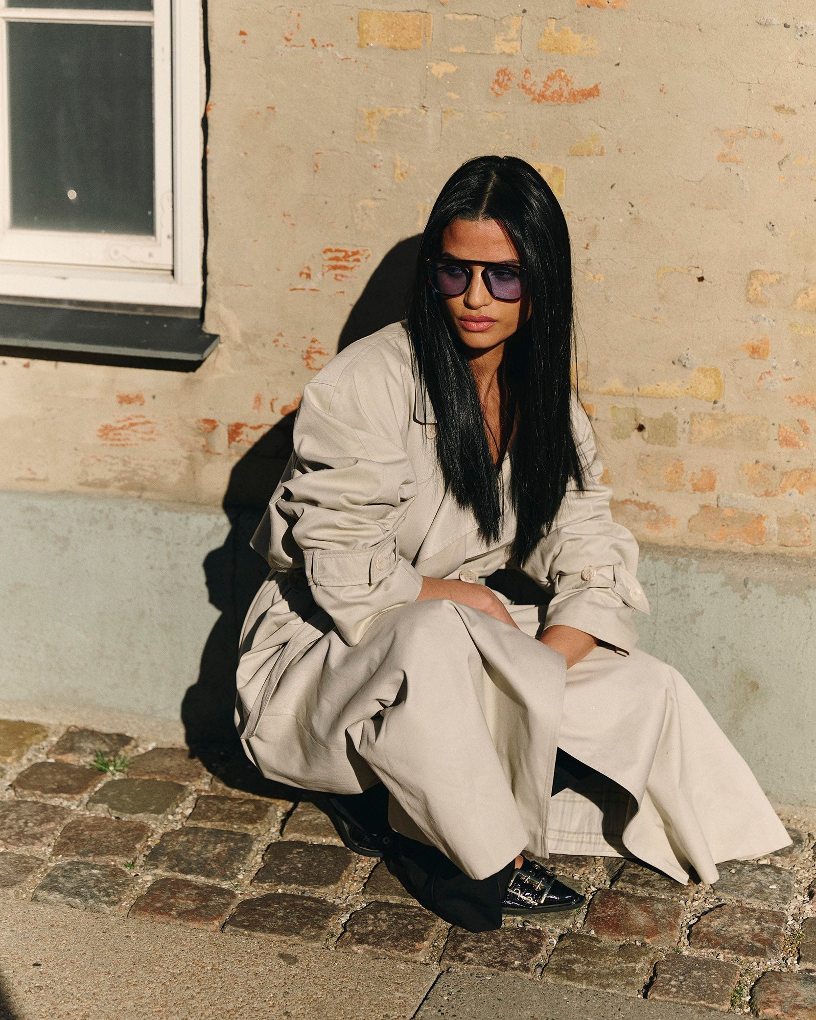 Woman in a black synthetic wig and a beige outfit sitting against a textured wall.