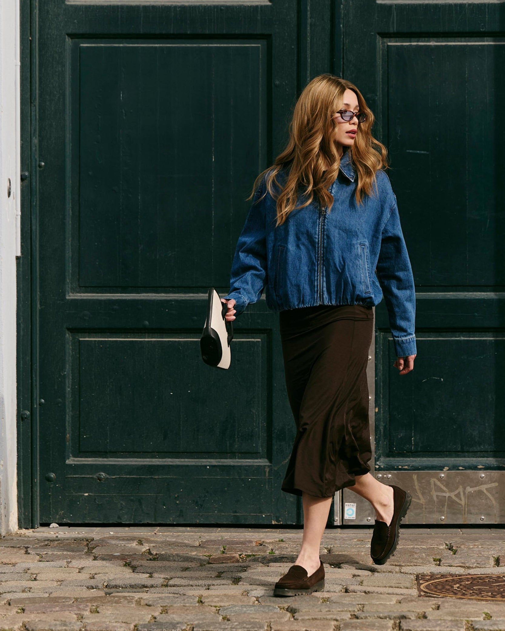 Woman in a brown balayage wig, blue jacket and black skirt standing in front of a large green door.