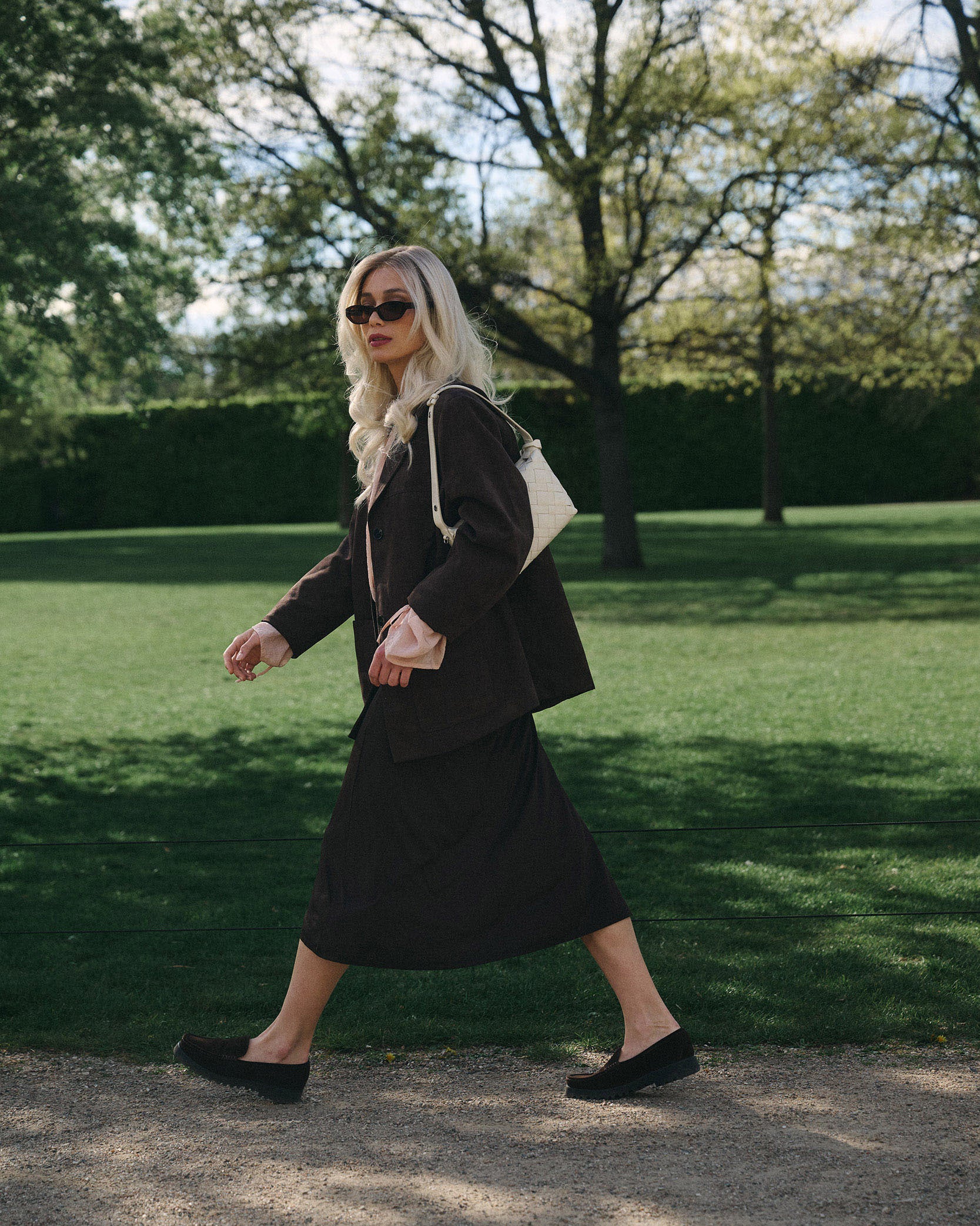 Woman in a blonde wig and  black suit walking outdoors on a grassy area with trees in the background