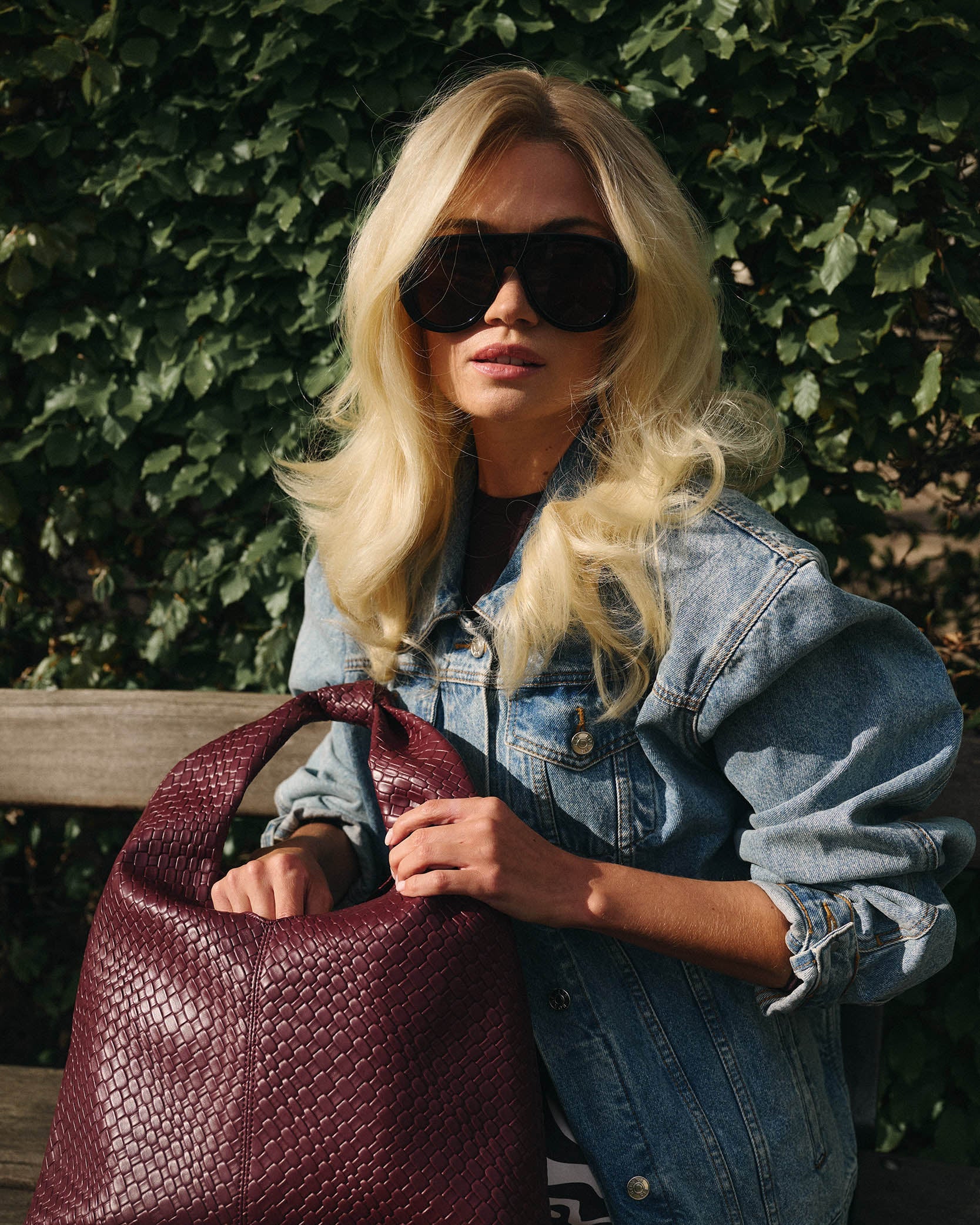 Woman in a blonde wig holding a maroon handbag wearing sunglasses and a denim jacket against a green leafy background.