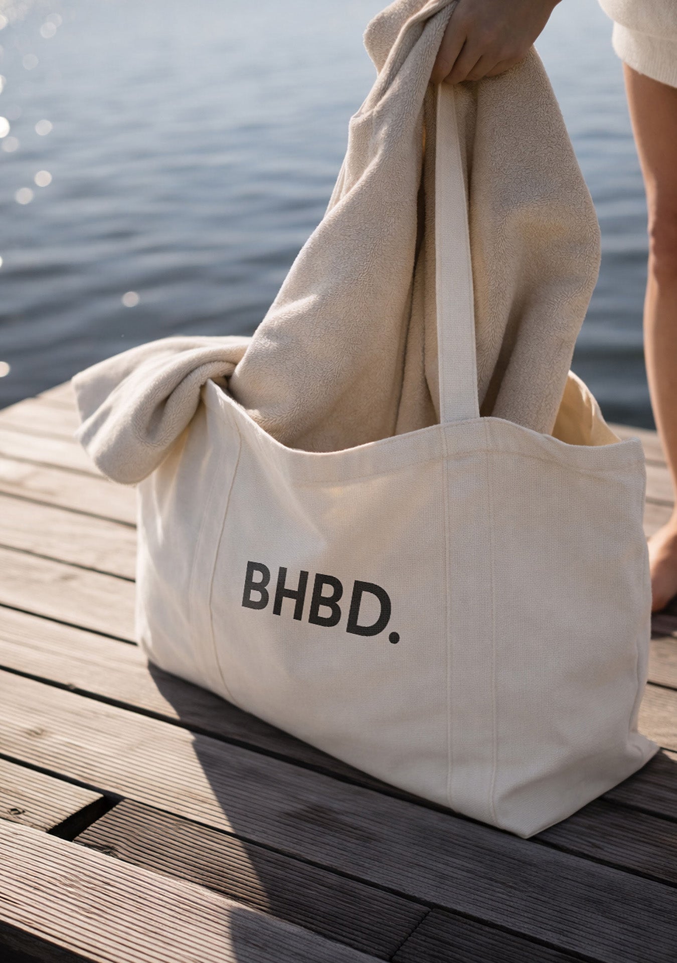 Beige tote bag with “BHBD.” logo on a wooden dock by the water, with a hand placing a towel inside.