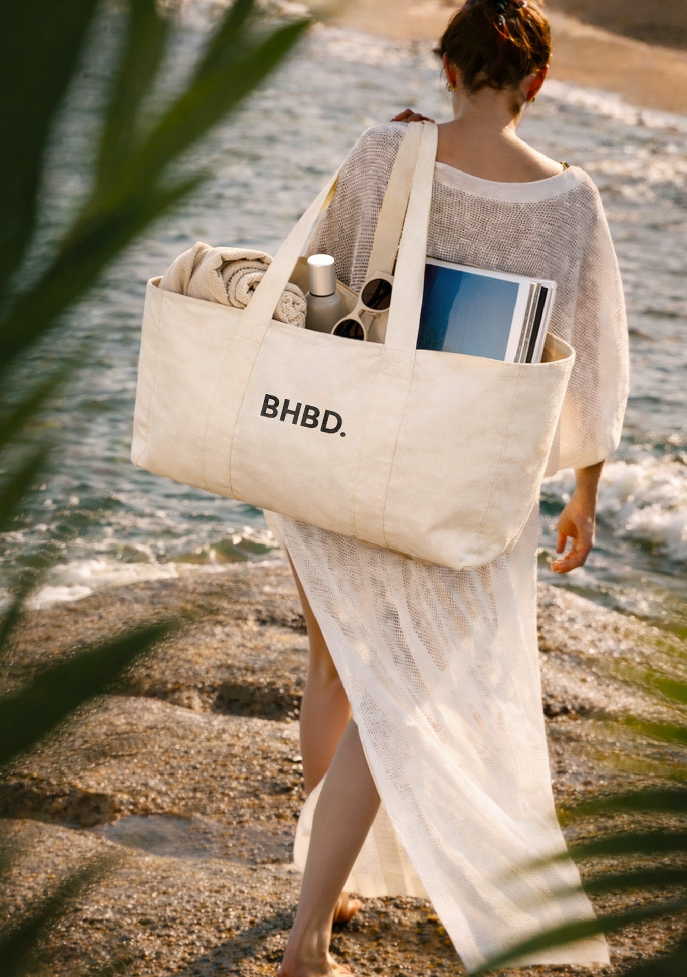 Woman walking along the shoreline carrying a beige “BHBD.” tote bag filled with a towel, bottle, sunglasses, and a magazine.