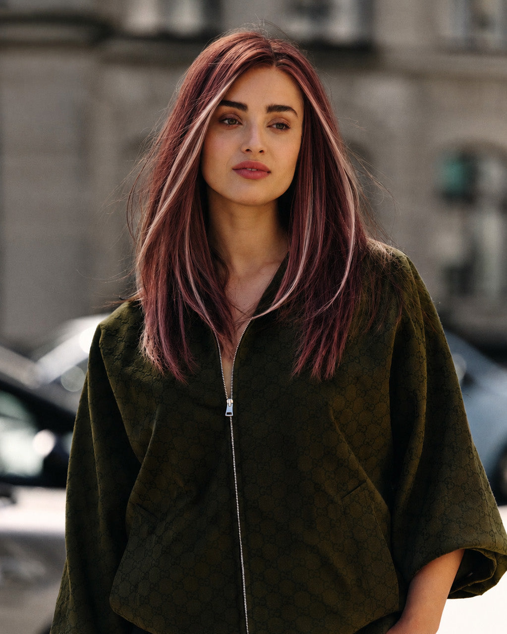 Woman with a plum-rose wig wearing a dark green jacket on a city street.