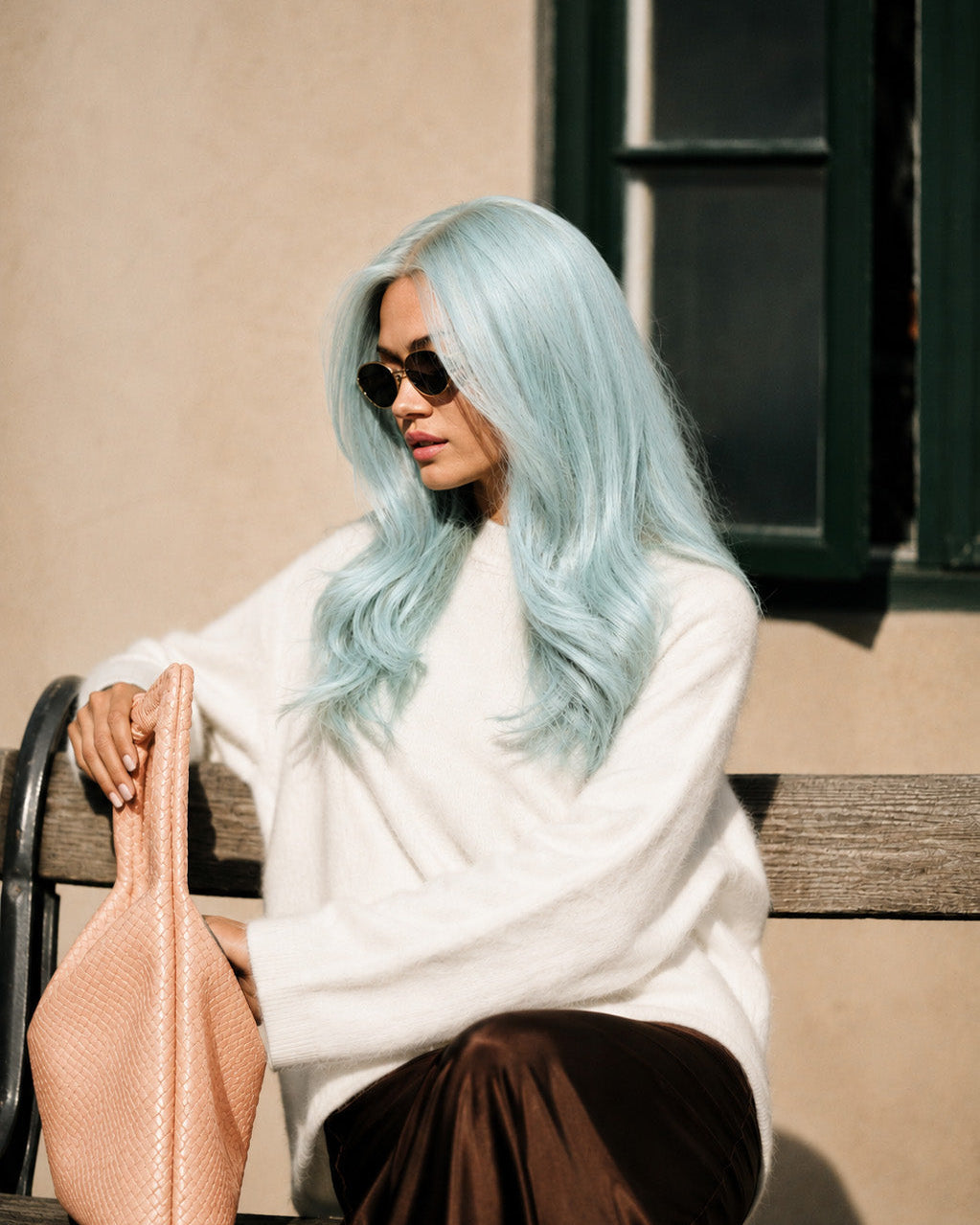 Person with light blue wig sitting on a bench wearing sunglasses and a white sweater.