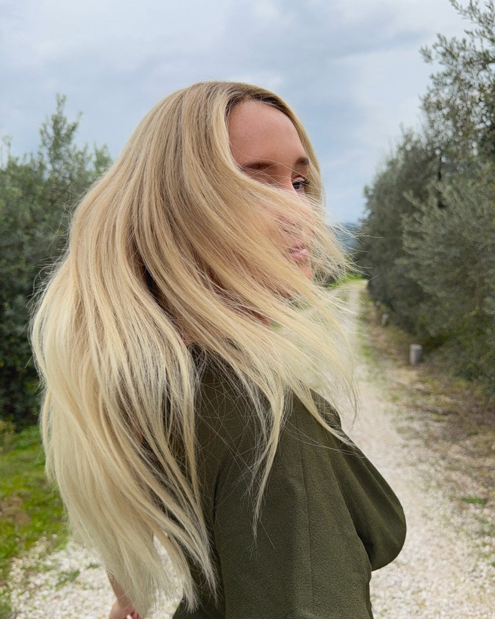 Vera Wig 40 cm light blonde with subtle golden hues and darker root, flowing naturally in daylight to show realistic shine and dimension, shown on model outdoors